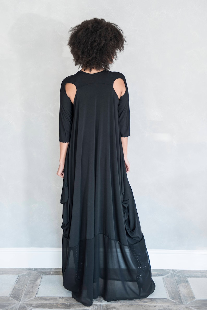 May include: A black floor-length dress featuring three-quarter length sleeves, shoulder cutouts, and a draped silhouette. The lower portion of the garment includes a sheer panel with subtle vertical detailing along the sides of the hemline.