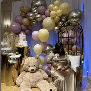 May include: A large teddy bear sits on the floor next to a gold metal birdcage chair. A multi-tiered cake and dessert stands are arranged on gold pedestals. An arch of balloons in shades of cream, mauve, and metallic gold fills the space.