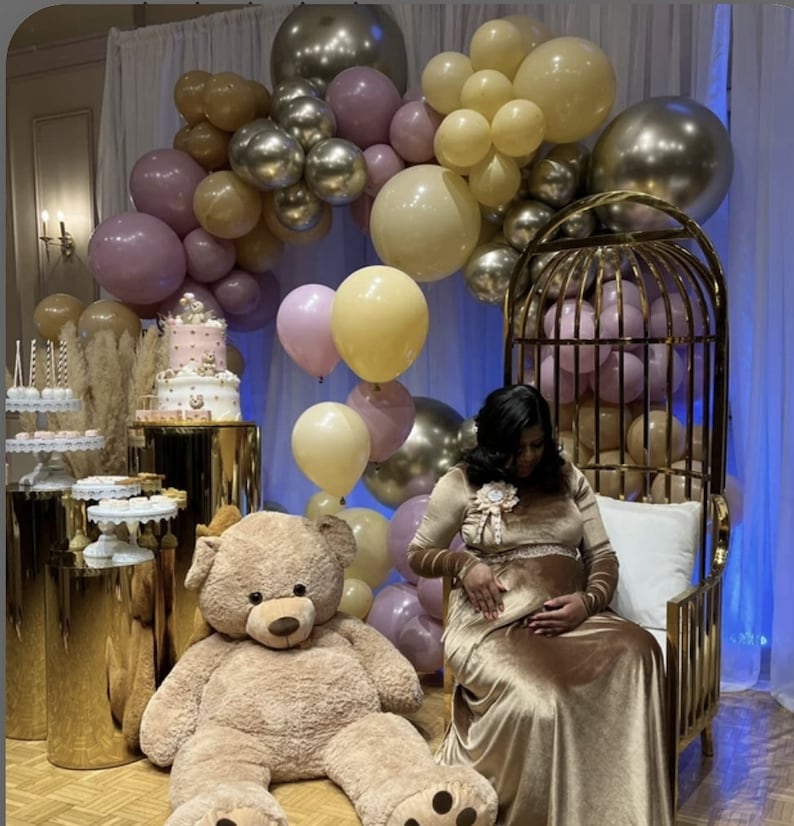 May include: A large teddy bear sits on the floor next to a gold metal birdcage chair. A multi-tiered cake and dessert stands are arranged on gold pedestals. An arch of balloons in shades of cream, mauve, and metallic gold fills the space.