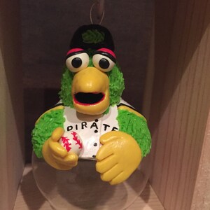 May include: A green and yellow bird wearing a baseball cap and jersey with the word "PIRATE" on it. The bird is holding a baseball and is inside a clear glass ornament.