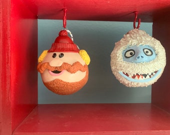 Puede incluir: Dos adornos navideños colgando de un estante rojo. El adorno de la izquierda es un hombre redondo, marrón, barbudo con un sombrero rojo y orejeras amarillas. El adorno de la derecha es un muñeco de nieve redondo, blanco y peludo con una cara azul y una sonrisa de dientes.