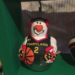 May include: A snowman wearing a black and yellow basketball jersey with the word "MARYLAND" and the number "2" on the front. The snowman is holding a basketball and wearing red and yellow mittens.