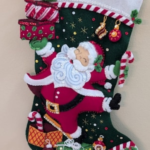 Könnte beinhalten: Ein dunkelgrüner Weihnachtsstrumpf aus Filz mit einer zentralen Weihnachtsmannfigur in rotem Anzug mit weißem Besatz. Der Strumpf ist mit applizierten Geschenkboxen, Zuckerstangen und kleinen Ornamenten verziert, die mit Pailletten bestickt sind.