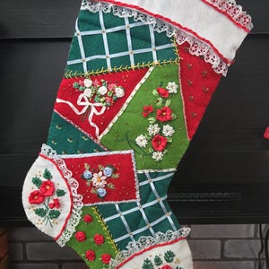 May include: A Christmas stocking with a patchwork design featuring red, green, and white panels. The stocking is adorned with embroidered floral details, white lace trim, and ribbon accents. The top and toe of the stocking are white.