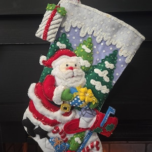 Puede incluir: Un calcetín navideño hecho a mano con Papá Noel, abetos cubiertos de nieve y un buzón. El calcetín está decorado con lentejuelas y aplicaciones de fieltro en rojo, blanco, verde y azul. También se incluye una caja de regalo y bastones de caramelo.
