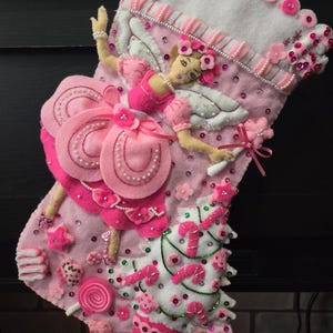 Könnte beinhalten: Ein rosa Weihnachtsstrumpf mit einem Feen-Design. Der Strumpf zeigt eine Filzfee mit Flügeln, einem rosa Kleid und einem floralen Kopfschmuck. Verzierungen sind rosa Perlen, Pailletten und ein Weihnachtsbaum mit Zuckerstangen.