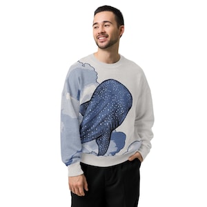 May include: A light gray crewneck sweatshirt featuring a large, dark blue whale shark graphic with white spots. The design includes stylized blue and white cloud-like patterns that extend across the front and onto the left sleeve of the garment.