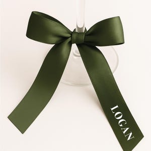Puede incluir: Una cinta de raso verde oscuro atada en un lazo alrededor del tallo de una copa de vino. La cinta tiene el nombre "LOGAN" impreso en blanco. El lazo está centrado, con las colas de la cinta extendiéndose hacia abajo.