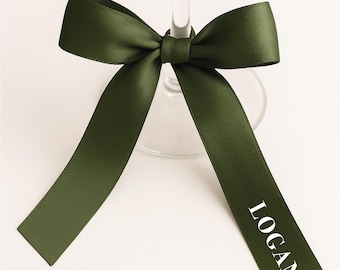 May include: A green satin ribbon tied in a bow around a wine glass stem. The ribbon has the name "LOGAN" printed in white. The bow is centered, with the ribbon tails extending downwards.