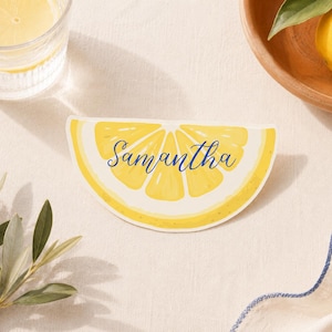Puede incluir: Una tarjeta de lugar con forma de limón con el nombre "Samantha" en escritura azul. La tarjeta está sobre una superficie blanca, con un vaso de agua y una rodaja de limón visibles. Un cuenco de madera con un limón y vegetación también están en la imagen.