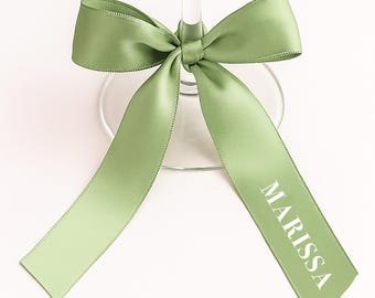 May include: A light green satin ribbon tied around a wine glass. The ribbon is knotted in a bow and has the name "MARISSA" printed in white. The glass is clear, and the background is white.