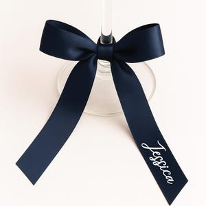 Op de afbeelding: Een marineblauw satijnen lint dat in een strik om de steel van een wijnglas is geknoopt. Op het lint staat de naam "Jessica" in een sierlijk lettertype. De strik is gecentreerd op het glas, met de uiteinden van het lint naar beneden hangend.