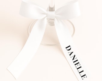 May include: A clear wine glass with a white ribbon tied around the stem. The ribbon has the name "DANIELLE" printed in black. The bow is centered, and the ribbon hangs down on either side of the glass.