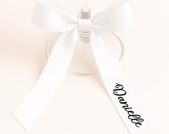 May include: A clear wine glass with a white satin ribbon tied around the stem. The ribbon has the name "Danielle" printed in black script. The background is a soft, neutral color.