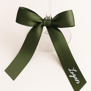 May include: A dark green satin ribbon tied in a bow around a wine glass stem. The ribbon has long tails, with the name "Logan" printed in white script on one tail. The bow is centered on the glass.