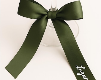 May include: A green satin ribbon tied in a bow around a wine glass stem. The ribbon has long tails and the name "Logan" is printed in white script on one tail. The bow is centered on a clear glass.