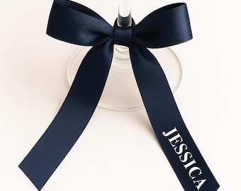 May include: A navy blue ribbon tied around a wine glass stem. The ribbon is knotted in a bow and has the name "JESSICA" printed in white. The glass sits on a white surface.