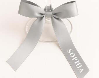 May include: A clear wine glass with a silver ribbon tied around the stem. The ribbon is in a bow shape, with the word "SOPHIA" printed in white on one of the ribbon tails. The background is white.