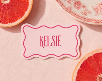 May include: A pink place card with a scalloped edge and the name "KELSIE" in dark pink lettering. The card is on a pink tablecloth, with grapefruit halves and a floral patterned plate in the background.