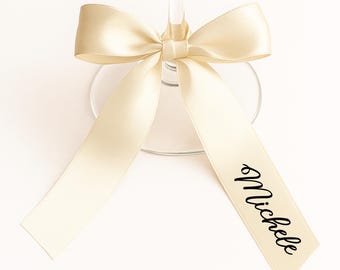 May include: A champagne-colored satin ribbon tied in a bow around a wine glass. The ribbon has the name "Michele" printed in black script. The glass is clear, and the background is white.