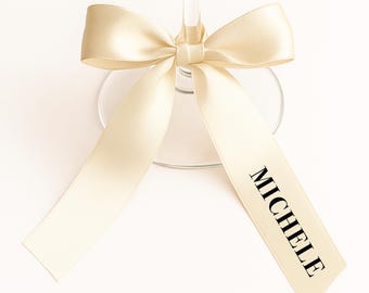 May include: A champagne-colored satin ribbon tied around a wine glass. The ribbon is knotted in a bow and has the name "MICHELE" printed in black. The glass is clear, and the background is white.