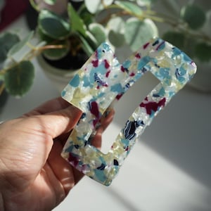 May include: A rectangular hair claw clip featuring a translucent base with a multi-colored confetti pattern in shades of light blue, white, yellow, and deep magenta. The accessory has a hollow center and a smooth, polished finish.