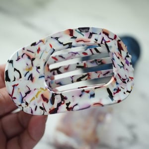 May include: A colorful hair claw clip with a rectangular shape and a series of horizontal bars. The clip features a speckled pattern with black, red, blue, and yellow accents on a white background. The clip is made of a glossy material.