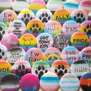 May include: A collection of colorful round buttons with various designs and text. Some buttons feature paw prints, rainbow stripes, and phrases like "Ew Gay," "Trans Rights are Human Rights," and "Pride." The buttons are a mix of colors.