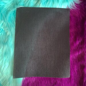 May include: A rectangular, dark gray textured fabric, possibly a mat or sheet, is centered in the image. The fabric has a fine, vertical ribbed pattern. The background features vibrant, fluffy faux fur in turquoise and deep purple.