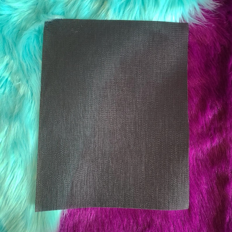 May include: A rectangular, dark gray textured fabric, possibly a mat or sheet, is centered in the image. The fabric has a fine, vertical ribbed pattern. The background features vibrant, fluffy faux fur in turquoise and deep purple.