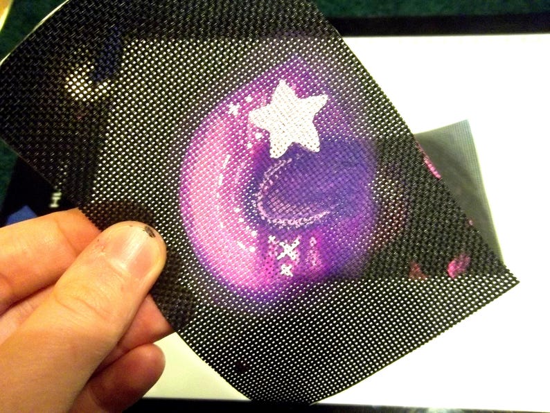 May include: A black mesh material with a purple and pink design featuring a white star and crescent moon. The mesh is held up, revealing the design underneath. The design appears to be a sticker or decal.