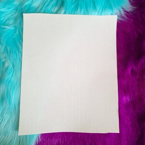 May include: A square, off-white piece of Aida cloth for cross-stitch projects. The fabric has a grid-like pattern of evenly spaced holes, ideal for needlework. The cloth is set against a backdrop of vibrant, fluffy turquoise and purple faux fur.