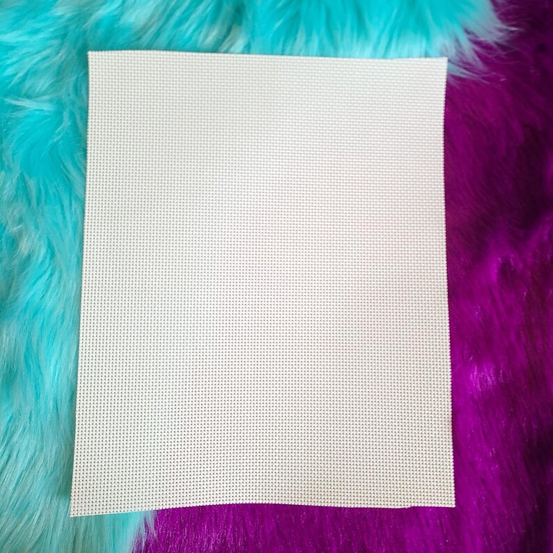 May include: A square, off-white piece of Aida cloth for cross-stitch projects. The fabric has a grid-like pattern of evenly spaced holes, ideal for needlework. The cloth is set against a backdrop of vibrant, fluffy turquoise and purple faux fur.
