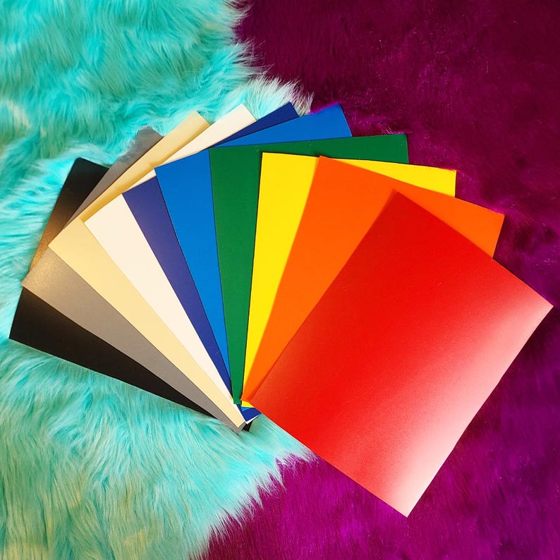 May include: A fan of colorful cardstock paper in various shades, including red, orange, yellow, green, blue, and black. The papers are arranged on a turquoise and purple faux fur background.