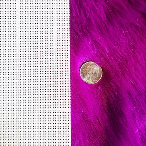 May include: A close-up shot features a dime coin on a vibrant, fuchsia-colored, faux fur surface. To the left, a white plastic canvas with evenly spaced square holes is visible. The coin is approximately 0.7 inches in diameter.
