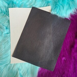 May include: Two rectangular sheets of craft material. One is off-white with a grid of small holes, the other is dark gray with a textured surface. The sheets are layered on a background of turquoise and purple faux fur.