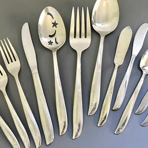 May include: A collection of vintage silver-toned flatware, including forks, knives, spoons, and a serving spoon with star and crescent cutouts. Each piece has a starburst design at the base of the handle. The arrangement is on a gray surface.