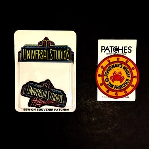 May include: Two sew-on souvenir patches. One patch is black and features the text "Universal Studios Hollywood". The other patch is red and yellow and features the text "Fisherman's Wharf San Francisco".