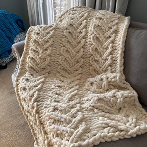 May include: A cream-colored, chunky knit blanket with a cable-knit design is draped over a gray sofa. The blanket's thick yarn creates a textured, cozy appearance. The blanket is folded over the sofa, showcasing its intricate pattern.