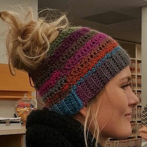 May include: A woman wearing a colorful striped crochet hat with a messy bun. The hat is made with a variety of colors, including pink, blue, green, and brown.