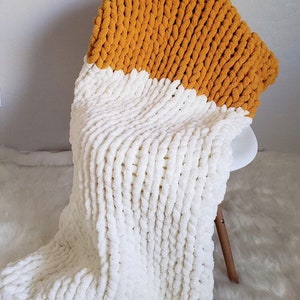 May include: A chunky knit throw blanket with a white bottom half and a mustard yellow top half. The blanket is draped over a chair.