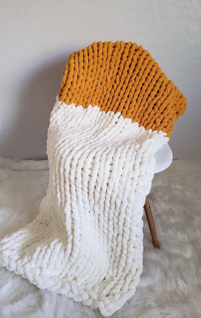 May include: A chunky knit throw blanket with a white bottom half and a mustard yellow top half. The blanket is draped over a chair.