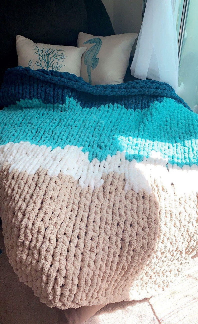 May include: A chunky knit throw blanket with stripes of teal, white, and beige. The blanket is draped over a couch with two decorative pillows featuring a seahorse and a branch design.