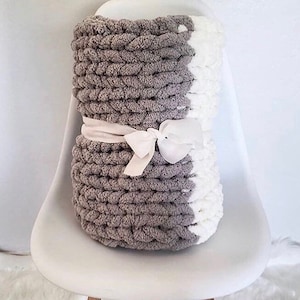 May include: A gray and white chunky knit throw blanket with a white satin ribbon tied around it. The blanket is folded and sitting on a white chair.