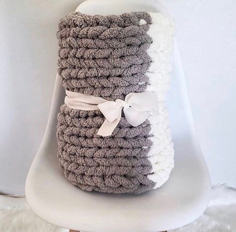 May include: A gray and white chunky knit throw blanket with a white satin ribbon tied around it. The blanket is folded and sitting on a white chair.