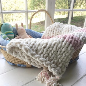 May include: A white and pink chunky knit baby blanket with tassels, in a wicker basket with blue and white gingham lining.