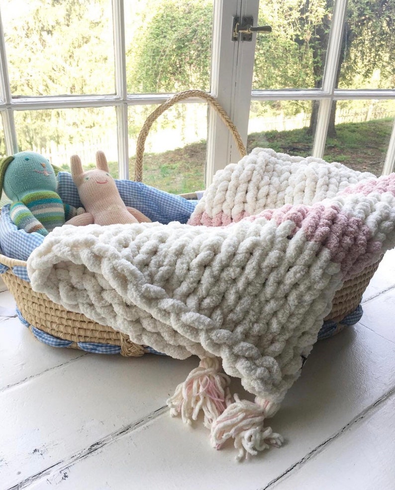 May include: A white and pink chunky knit baby blanket with tassels, in a wicker basket with blue and white gingham lining.
