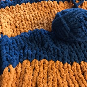 May include: A chunky knit blanket with alternating stripes of mustard yellow and dark blue yarn. A ball of dark blue yarn is visible in the upper right corner of the image.