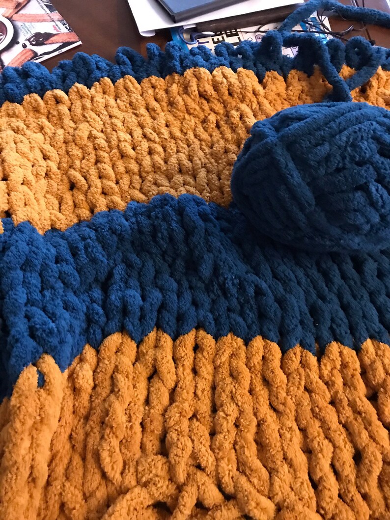 May include: A chunky knit blanket with alternating stripes of mustard yellow and dark blue yarn. A ball of dark blue yarn is visible in the upper right corner of the image.