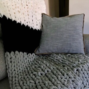 May include: A gray chunky knit throw blanket with a white and black patterned throw pillow and a gray throw pillow with a brown border on a gray couch.